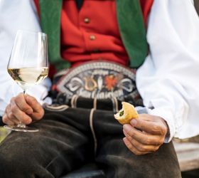 Ein Mann in traditioneller Kleidung hält ein Glas Weißwein und ein Stück Fingerfood in den Händen