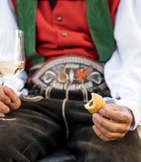 Ein Mann in traditioneller Kleidung hält ein Glas Weißwein und ein Stück Fingerfood in den Händen