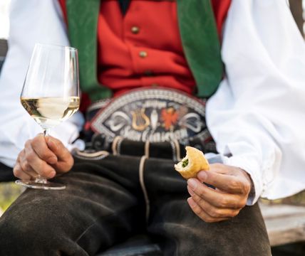 A man wearing traditional clothes is holding a glass of white wine and a piece of finger food