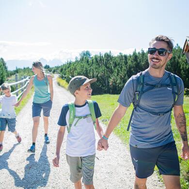 Lachende Familie beim Wandern auf einem sonnigen Weg mit Holzzaun und grüner Landschaft.