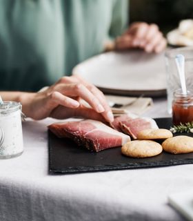 Eine Person greift nach einer Scheibe Speck auf einer Servierplatte mit Südtiroler Spezialitäten wie Käse, Brot, Kräuterquark und Marmelade.