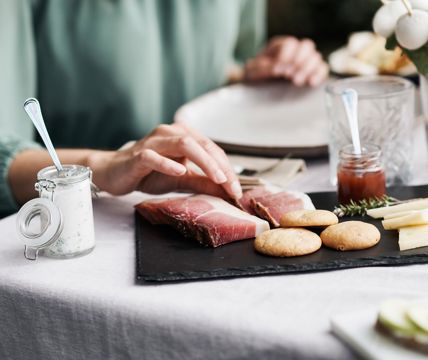 Eine Person greift nach einer Scheibe Speck auf einer Servierplatte mit Südtiroler Spezialitäten wie Käse, Brot, Kräuterquark und Marmelade.