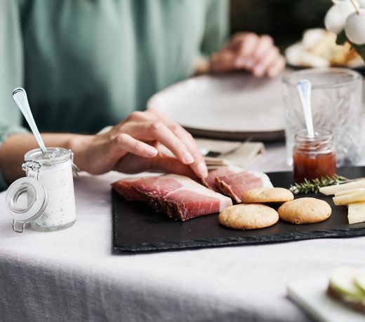 A person reaches for a slice of speck on a serving board with South Tyrolean specialties like cheese, bread, herb quark, and jam.