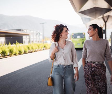 Two women are chatting and smiling at a modern bus stop, with a green bus in the background.