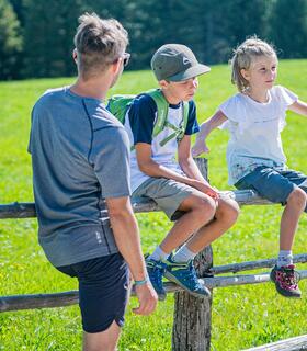 A family takes a break during a hike and enjoys the sunshine on a meadow with a wooden fence.
