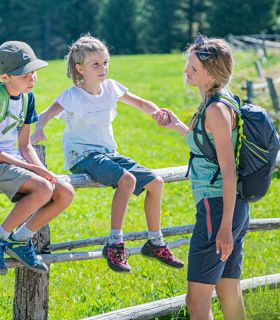 A family takes a break during a hike and enjoys the sunshine on a meadow with a wooden fence.