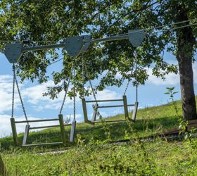 Zwei Schaukeln aus Metall hängen an einem Seilsystem auf einer Wiese unter Bäumen.