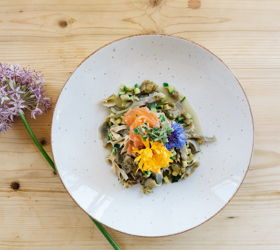 Teller mit Pasta, Gemüse, Lachs und essbaren Blüten, serviert auf einem hellem Holztisch mit einer Blume.