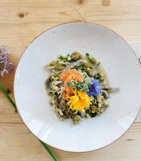 Plate of pasta with vegetables, salmon and edible flowers on light wooden table with a flower.