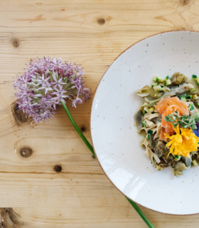 Plate of pasta with vegetables, salmon and edible flowers on light wooden table with a flower.