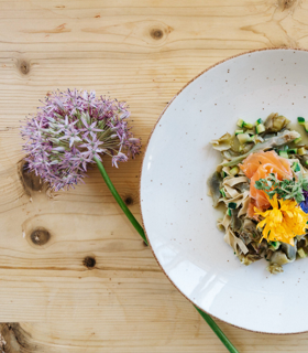 Teller mit Pasta, Gemüse, Lachs und essbaren Blüten, serviert auf einem hellem Holztisch mit einer Blume.
