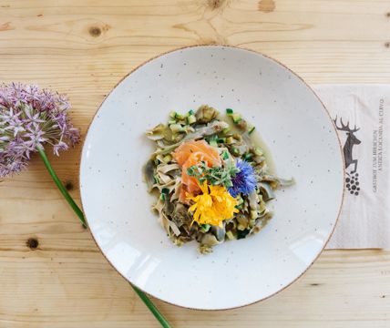Teller mit Pasta, Gemüse, Lachs und essbaren Blüten, serviert auf einem hellem Holztisch mit einer Blume.