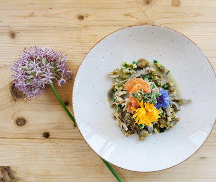 Teller mit Pasta, Gemüse, Lachs und essbaren Blüten, serviert auf einem hellem Holztisch mit einer Blume.